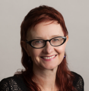 Headshot of Dr. Monica Noy. A woman with long red hair, wearing thick, black, cat-eye glasses and a black knit shirt.